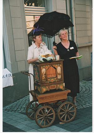 Leierkasten Zwei unserer freundlichen Mitarbeiterinnen bedienen unter einem Regenschirm einen schönen, alten Leierkasten.